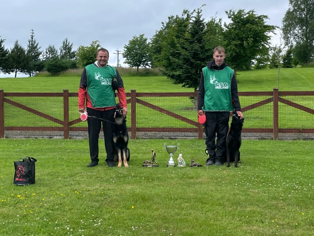 IMG-20250808-WA0076 Handlers David and Raymond posing with their trophies at the Irish Nationals while their dogs, Jagger and Deadly, watch attentively, showcasing focus and championship-level training.